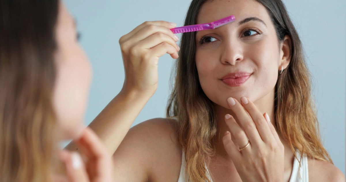 A woman in Ventura, CA looking into a mirror while shaving her eyebrow with a pink razor, highlighting Dermaplaning.